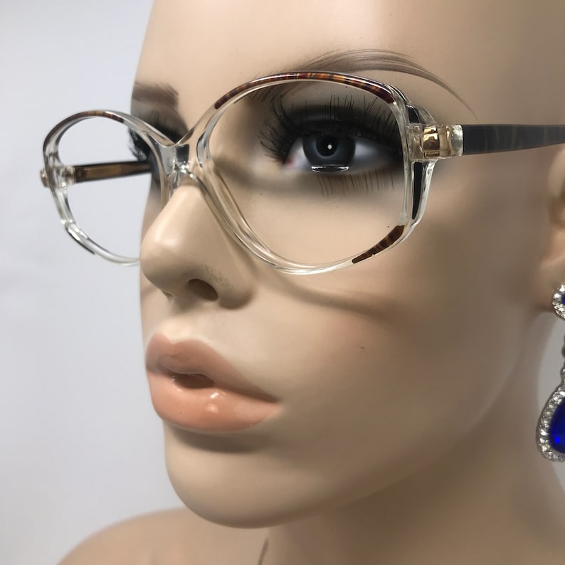 May include: Clear-framed eyeglasses with a rounded square shape and brown tortoiseshell detailing on the top rims and temples. The frames feature gold-coloured accents at the hinges. The glasses are displayed on a mannequin.