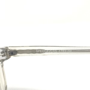 May include: Close-up of a clear eyeglasses frame arm. The text "SPECSAVERS 1753156" is printed in a dark gray color on the arm. The frame is made of a transparent material and is set against a white background.