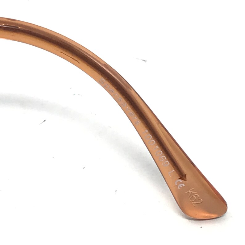 K&ouml;nnte beinhalten: Nahaufnahme des braunen Kunststoffarms einer Brille. Der Arm ist mit dem Text "Eyeglasses 100-0501 CE K62" beschriftet.