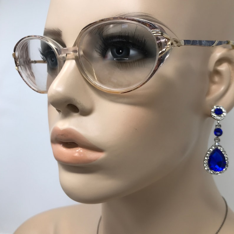 May include: Vintage eyeglasses with a light brown frame and clear lenses. The glasses have a rounded oval shape. A pair of blue and silver drop earrings are visible on the right side of the image.