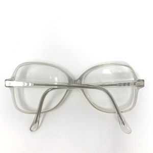 May include: Clear, oversized eyeglasses with a light gray frame. The glasses have a rectangular shape with rounded edges and a bridge connecting the two lenses. The temples curve down to the ear pieces. The glasses are viewed from the back.