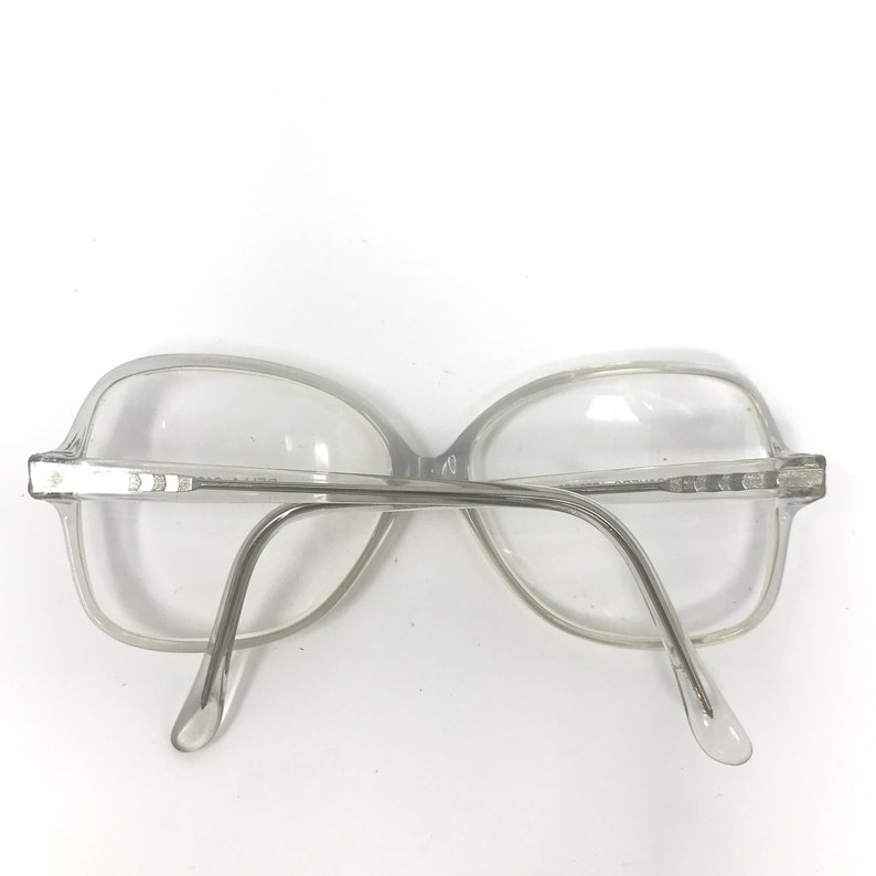 May include: Clear, oversized eyeglasses with a light gray frame. The glasses have a rectangular shape with rounded edges and a bridge connecting the two lenses. The temples curve down to the ear pieces. The glasses are viewed from the back.
