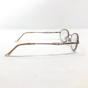 May include: A pair of eyeglasses with a thin, bronze-colored metal frame and oval lenses. The temples are a light tan color and curve gently. The frame features a decorative heart-shaped design near the hinges. The glasses are set against a white background.