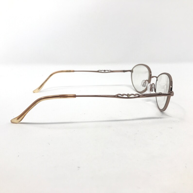 May include: A pair of eyeglasses with a thin, bronze-colored metal frame and oval lenses. The temples are a light tan color and curve gently. The frame features a decorative heart-shaped design near the hinges. The glasses are set against a white background.