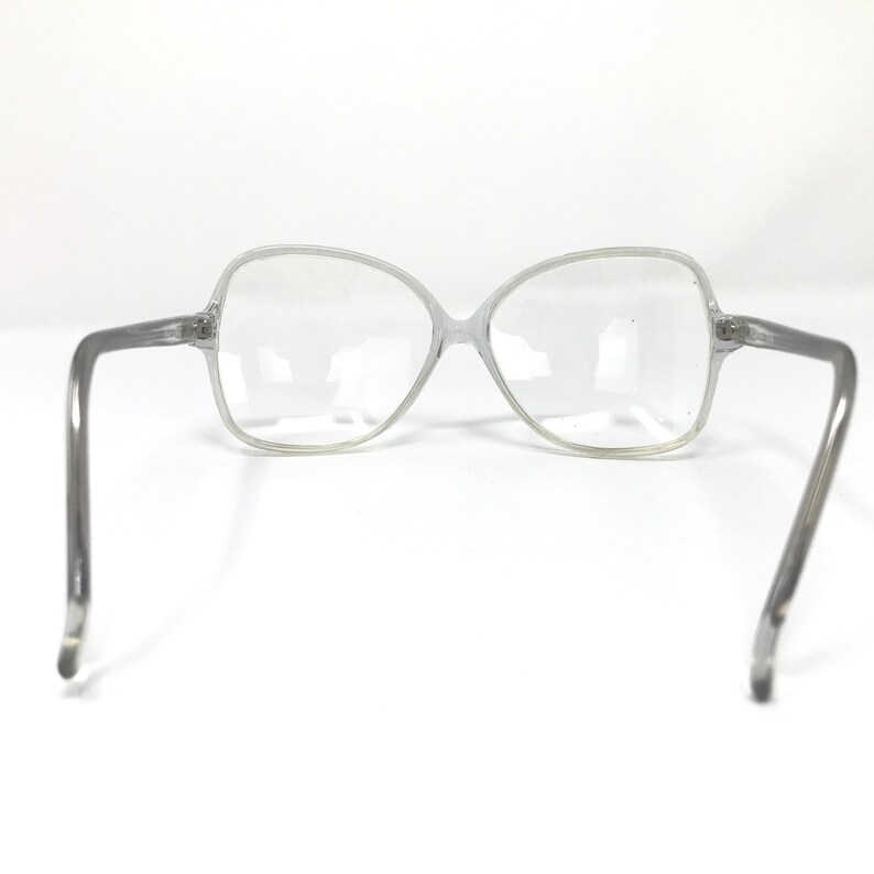 May include: Clear, oversized eyeglasses with a light gray frame. The glasses have a butterfly shape and are set against a white background. The temples are straight and extend to the sides.