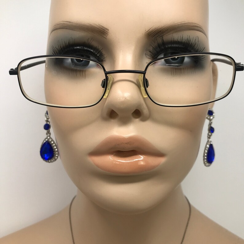 Puede incluir: Gafas rectangulares negras con montura fina y lentes transparentes. La imagen tambi&eacute;n presenta un par de pendientes colgantes con engaste plateado y grandes piedras preciosas azules en forma de l&aacute;grima. Los pendientes est&aacute;n acentuados con piedras azules m&aacute;s peque&ntilde;as.