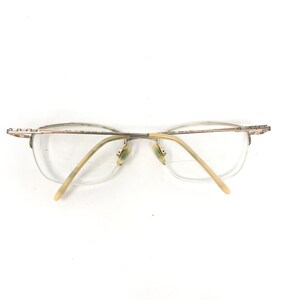 May include: A pair of eyeglasses with a gold-colored metal frame and clear lenses. The glasses have a half-rim design, with the frame extending along the top and sides of the lenses. The temples are a light beige color.