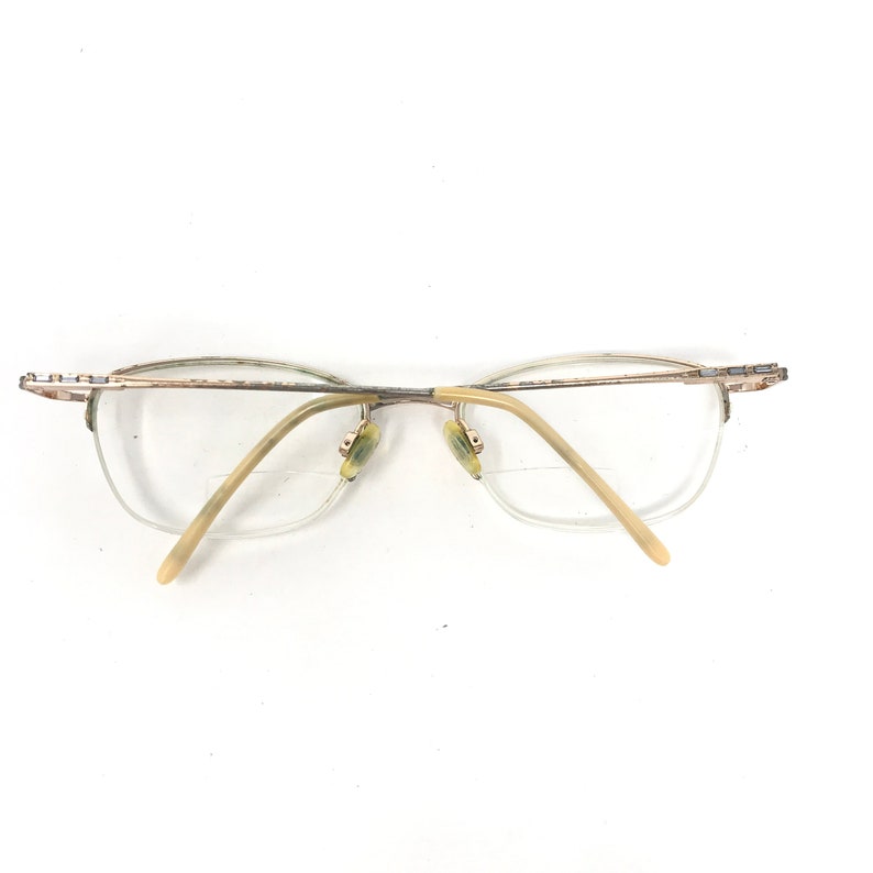 May include: A pair of eyeglasses with a gold-colored metal frame and clear lenses. The glasses have a half-rim design, with the frame extending along the top and sides of the lenses. The temples are a light beige color.