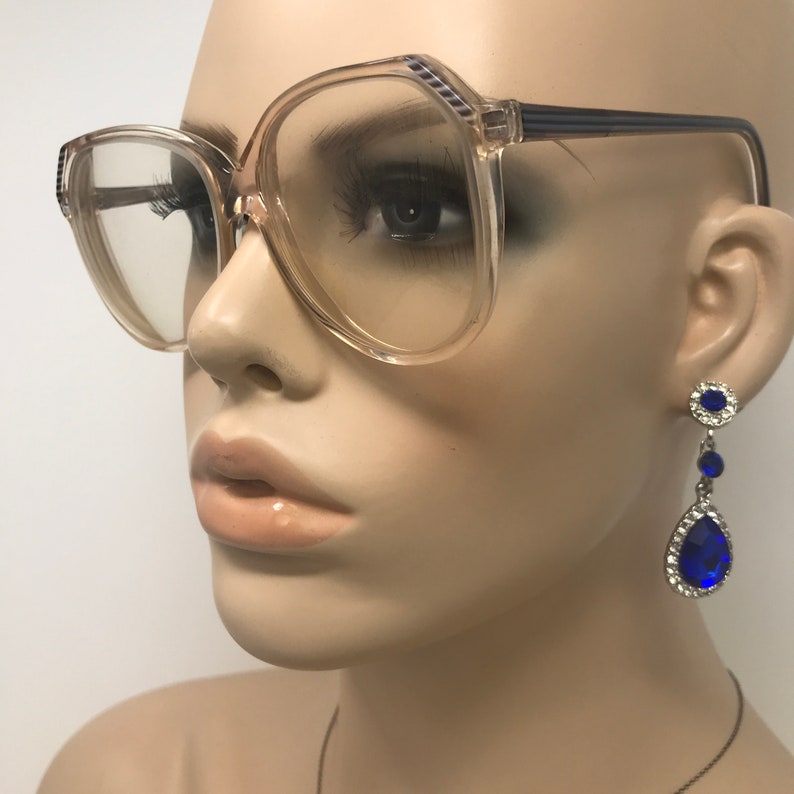 May include: Clear-lensed eyeglasses with a light brown frame and dark blue accents. The eyeglasses are paired with dangling blue and silver earrings. The earrings have a teardrop shape with a large blue stone.