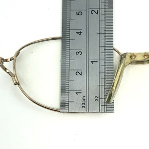 Peut inclure: Une monture de lunettes en m&eacute;tal dor&eacute; avec un pont de nez et des embouts de branches en plastique transparent. La monture est mesur&eacute;e &agrave; l'aide d'une r&egrave;gle, montrant qu'elle mesure environ 2,5 cm de large.