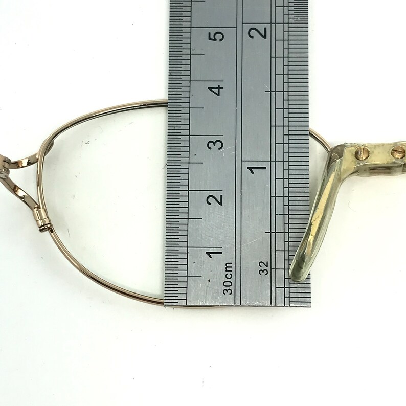 Peut inclure: Une monture de lunettes en m&eacute;tal dor&eacute; avec un pont de nez et des embouts de branches en plastique transparent. La monture est mesur&eacute;e &agrave; l'aide d'une r&egrave;gle, montrant qu'elle mesure environ 2,5 cm de large.