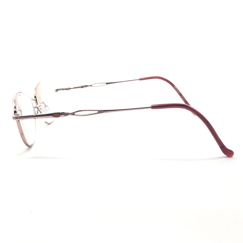 Puede incluir: Gafas con una fina montura de metal plateado y lentes ovaladas. Las patillas son de un color burdeos intenso, curv&aacute;ndose suavemente para descansar detr&aacute;s de las orejas. El dise&ntilde;o es minimalista y elegante.