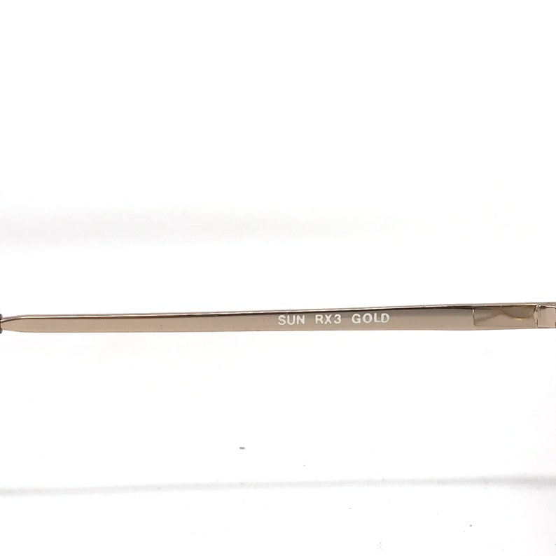May include: Close-up of a gold-colored eyeglass frame arm. The text "SUN RX3 GOLD" is printed in a dark font along the arm. The frame is thin and appears to be made of metal.