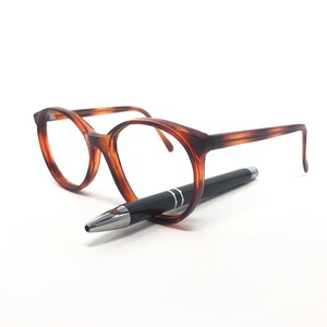 May include: A pair of tortoiseshell eyeglasses with a black pen resting beneath them. The glasses have a round frame and the pen has silver accents.