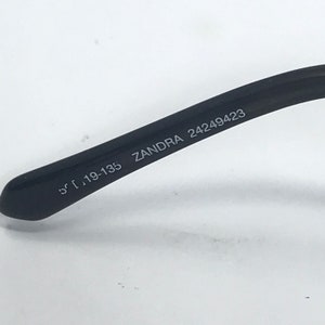May include: Close-up of the black arm of a pair of glasses with the text "ZANDRA 24249423" and "51 19-135" printed on it.
