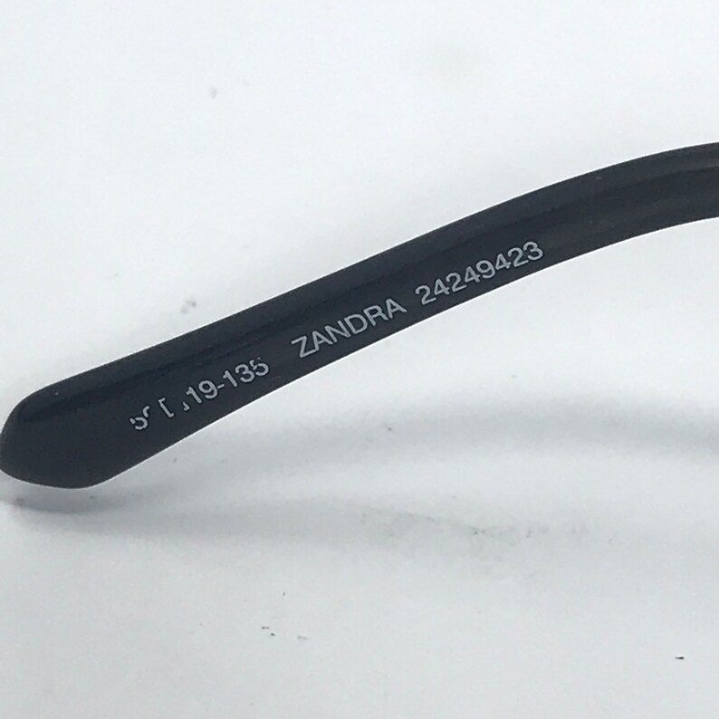 May include: Close-up of the black arm of a pair of glasses with the text "ZANDRA 24249423" and "51 19-135" printed on it.