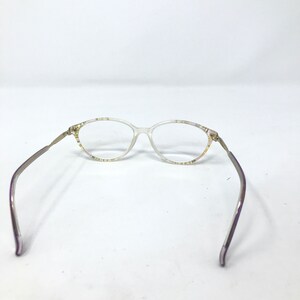 May include: Clear oval eyeglasses with thin, silver-toned arms and a decorative pattern on the frame. The glasses are set against a white background, showcasing their delicate design and vintage aesthetic.