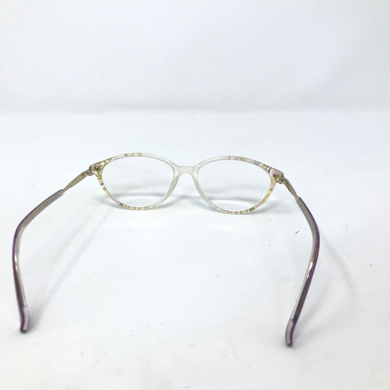 May include: Clear oval eyeglasses with thin, silver-toned arms and a decorative pattern on the frame. The glasses are set against a white background, showcasing their delicate design and vintage aesthetic.