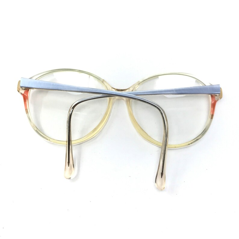 Puede incluir: Un par de gafas de pl&aacute;stico transparente con un puente de metal plateado y detalles de color rosa claro en las patillas.