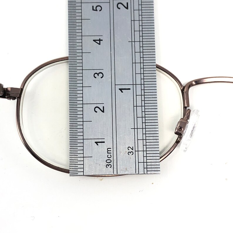 Puede incluir: Primer plano de gafas con montura met&aacute;lica. Se coloca una regla sobre la lente para medir el di&aacute;metro. Las gafas tienen una forma redonda cl&aacute;sica y lentes transparentes. La montura es de color met&aacute;lico oscuro.