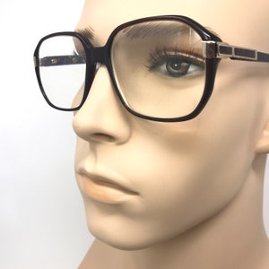 May include: Dark brown square-framed spectacles with clear lenses. The spectacles have a thick frame with a rectangular shape and silver accents on the temples. The spectacles are displayed on a mannequin.