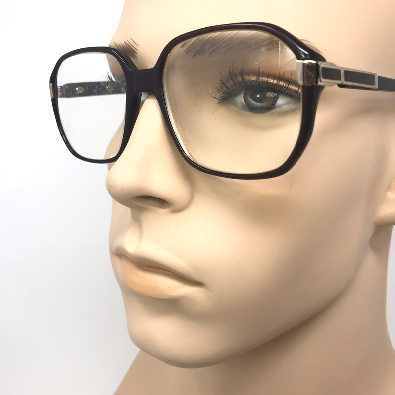 May include: Dark brown square-framed spectacles with clear lenses. The spectacles have a thick frame with a rectangular shape and silver accents on the temples. The spectacles are displayed on a mannequin.