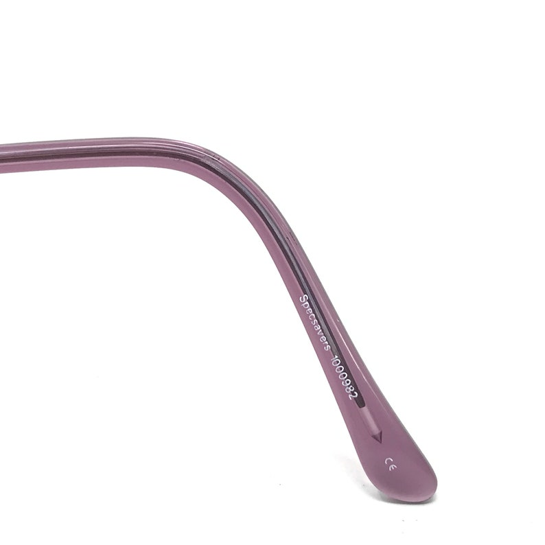 Puede incluir: Primer plano de la patilla de unas gafas. La patilla es de color morado apagado y presenta el texto "Specsavers 1000982" impreso en blanco. El extremo de la patilla tiene forma redondeada.
