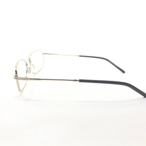 May include: A pair of eyeglasses with a silver metal frame and black temples. The lenses are clear, and the glasses are set against a white background. The design is minimalist and modern.