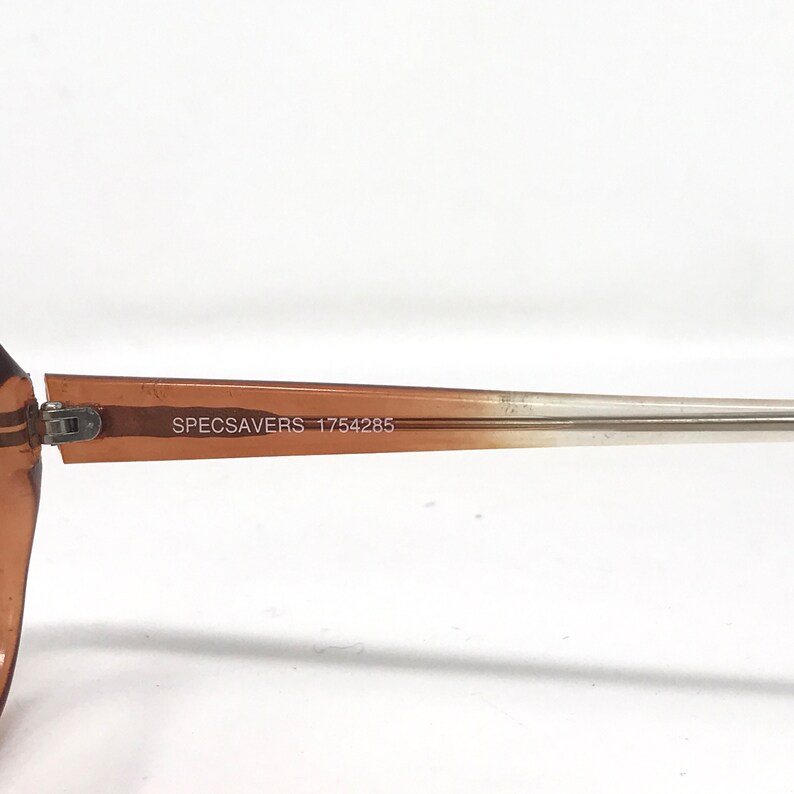 May include: Close-up of a pair of eyeglasses. The frame is a gradient of brown and silver, with the text "SPECSAVERS 1754285" printed on the temple arm. The glasses are set against a white background.