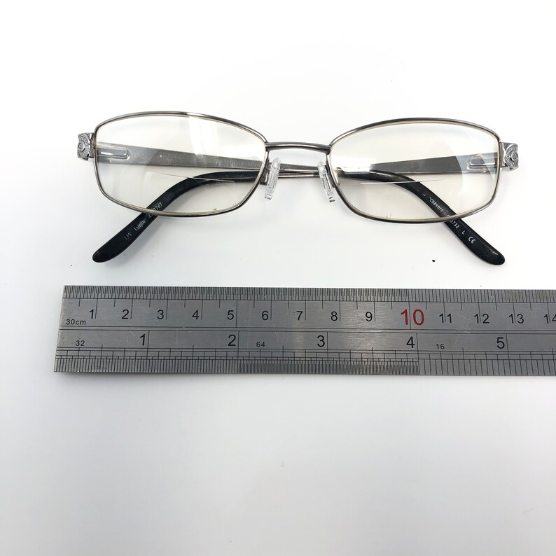 May include: Silver-framed eyeglasses with clear lenses. The glasses have a rectangular shape and black temples. A ruler is placed below the glasses, showing measurements in inches. The glasses are on a white surface.