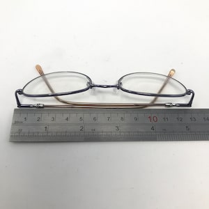 May include: A pair of eyeglasses with oval lenses and a dark blue frame. The temples are a light brown color. A ruler is placed beneath the glasses, showing a length of approximately 5 inches.