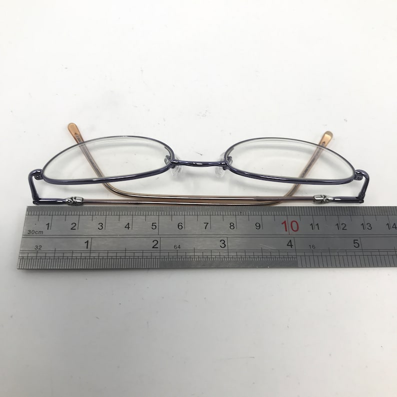 May include: A pair of eyeglasses with oval lenses and a dark blue frame. The temples are a light brown color. A ruler is placed beneath the glasses, showing a length of approximately 5 inches.