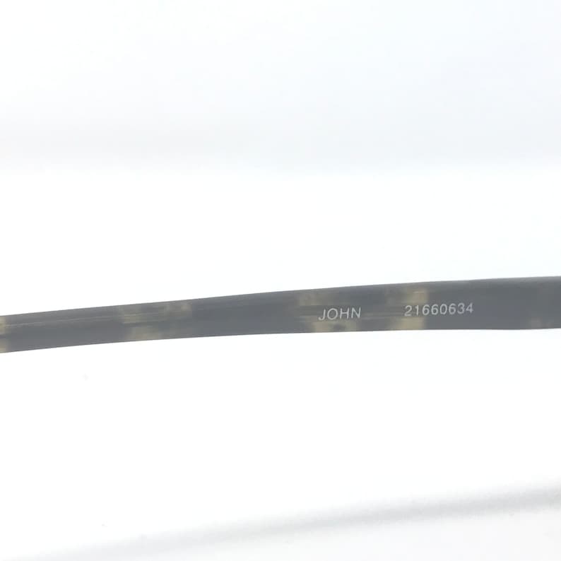 Puede incluir: Una secci&oacute;n de un par de gafas con un patr&oacute;n moteado gris y marr&oacute;n. La palabra "JOHN" est&aacute; impresa en la patilla, seguida del n&uacute;mero "21660634".