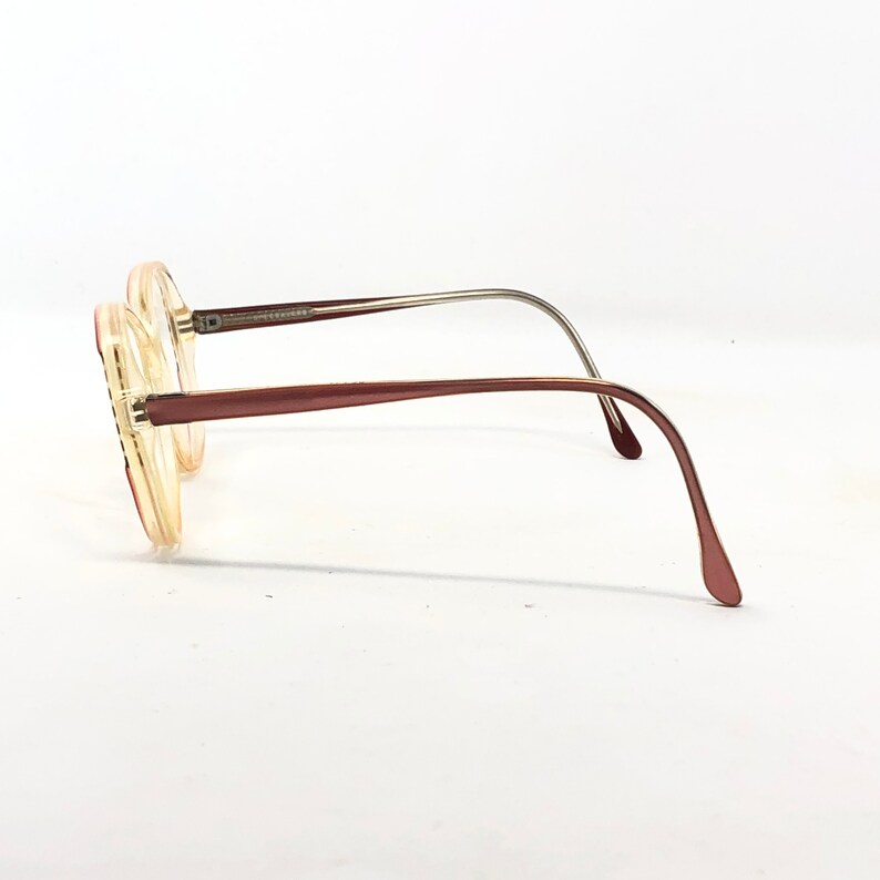 Puede incluir: Un par de gafas vintage con montura y patillas de color marr&oacute;n claro. Las lentes son de color amarillo claro. Las patillas son de color marr&oacute;n rojizo oscuro con detalles met&aacute;licos plateados. Las gafas est&aacute;n sobre un fondo blanco.
