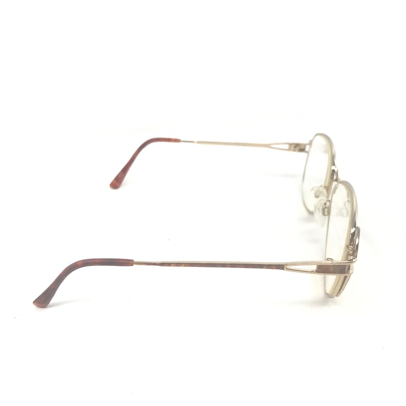 K&ouml;nnte beinhalten: Eine Brille mit einem goldfarbenen Metallrahmen und braunen B&uuml;geln. Die Gl&auml;ser sind klar, und die Brille ist vor einem wei&szlig;en Hintergrund platziert. Das Design ist eine klassische, runde Form.