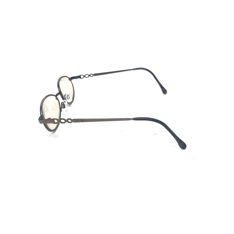 May include: Oval-shaped eyeglasses with a dark brown metal frame. The temples feature a decorative chain-link design. The lenses are a light tint, and the earpieces are black. The glasses are set against a white background.