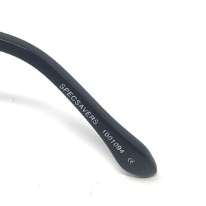 Puede incluir: Primer plano de una patilla de gafas negra. El texto "SPECSAVERS 1001094 CE" est&aacute; impreso en blanco en la patilla. La patilla est&aacute; en &aacute;ngulo y parece estar hecha de un material liso y mate.