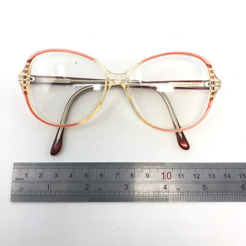 Puede incluir: Gafas con montura transparente con un acento rojo y amarillo en el borde superior y las patillas. Las gafas tienen un puente nasal y patillas marrones. Una regla de metal est&aacute; colocada debajo.