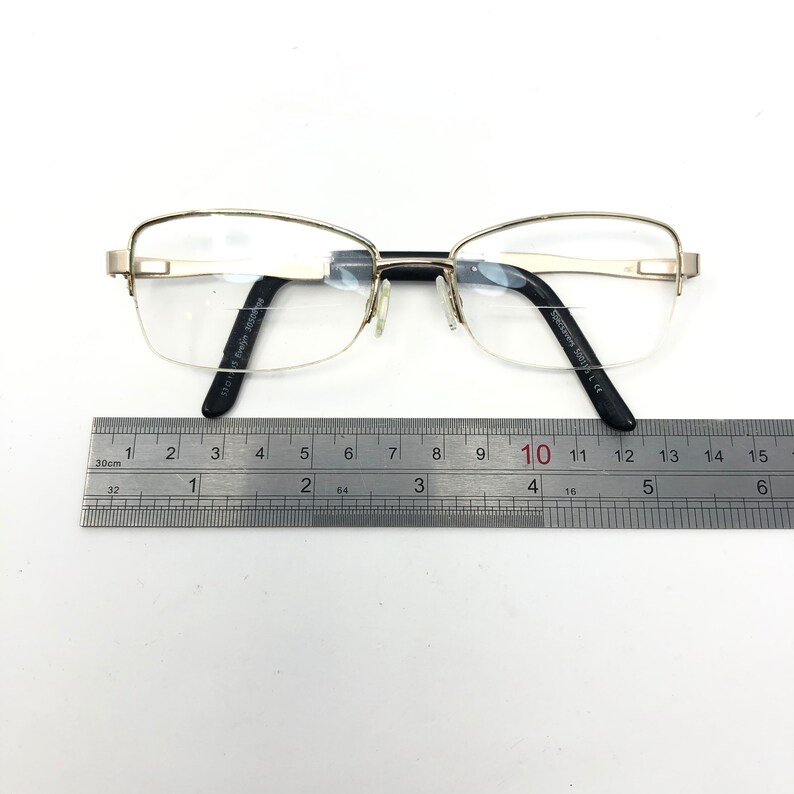 Pu&ograve; includere: Occhiali argentati con montatura rettangolare e lenti trasparenti. Le aste nere riportano la scritta "Evelyn 300012". Un righello misura gli occhiali, circa 15 cm di lunghezza.