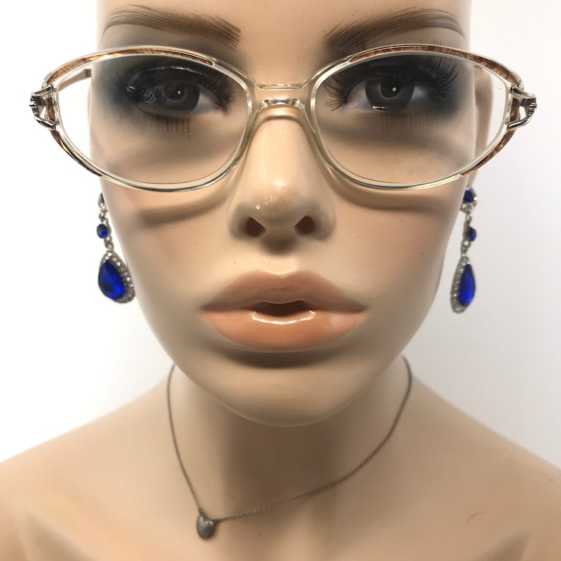 K&ouml;nnte beinhalten: Brille mit transparentem Rahmen und braun gemusterter Oberkante. Die Brille befindet sich auf einem Schaufensterpuppenkopf, der auch blaue Tr&auml;nenohrringe und eine silberne Halskette mit Herzanh&auml;nger tr&auml;gt.