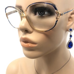 May include: Clear-framed eyeglasses with blue and gold accents. The glasses are paired with a pair of blue teardrop earrings with a silver chain. The earrings have a round top and a large teardrop-shaped blue gem.