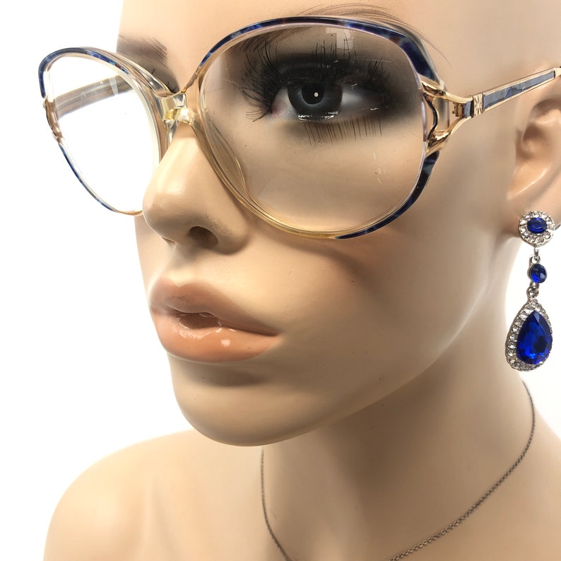 May include: Clear-framed eyeglasses with blue and gold accents. The glasses are paired with a pair of blue teardrop earrings with a silver chain. The earrings have a round top and a large teardrop-shaped blue gem.