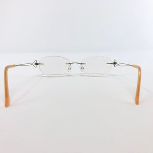 May include: Rimless eyeglasses with oval lenses and thin, silver-toned metal frames. The temples are a light tan color. The glasses are set against a white background.