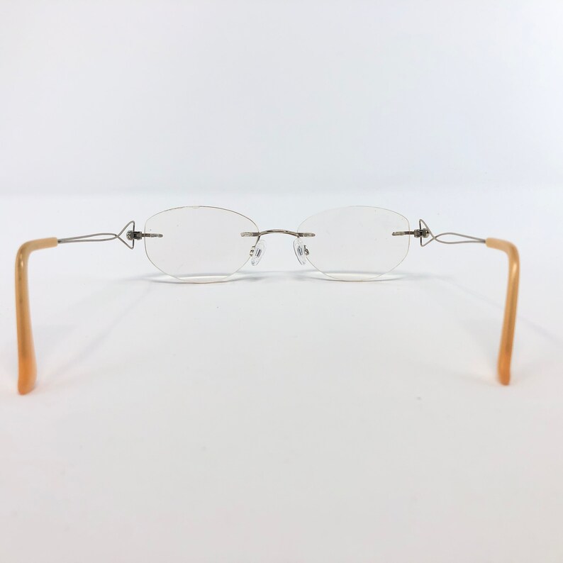 May include: Rimless eyeglasses with oval lenses and thin, silver-toned metal frames. The temples are a light tan color. The glasses are set against a white background.