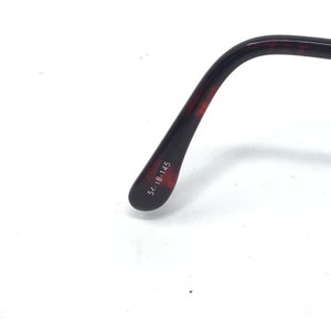 May include: Close-up of the temple arm of eyeglasses. The arm is a dark brown with a tortoiseshell pattern. The size is printed on the arm: 54-18-145.