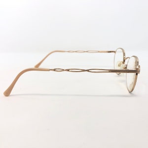 K&ouml;nnte beinhalten: Goldfarbene Brille mit zartem, ovalem Rahmen und einem einzigartigen, ineinander verschlungenen Design an den B&uuml;geln. Die B&uuml;gel und Ohrauflagen sind hellbeige. Die Gl&auml;ser sind klar, vor wei&szlig;em Hintergrund.