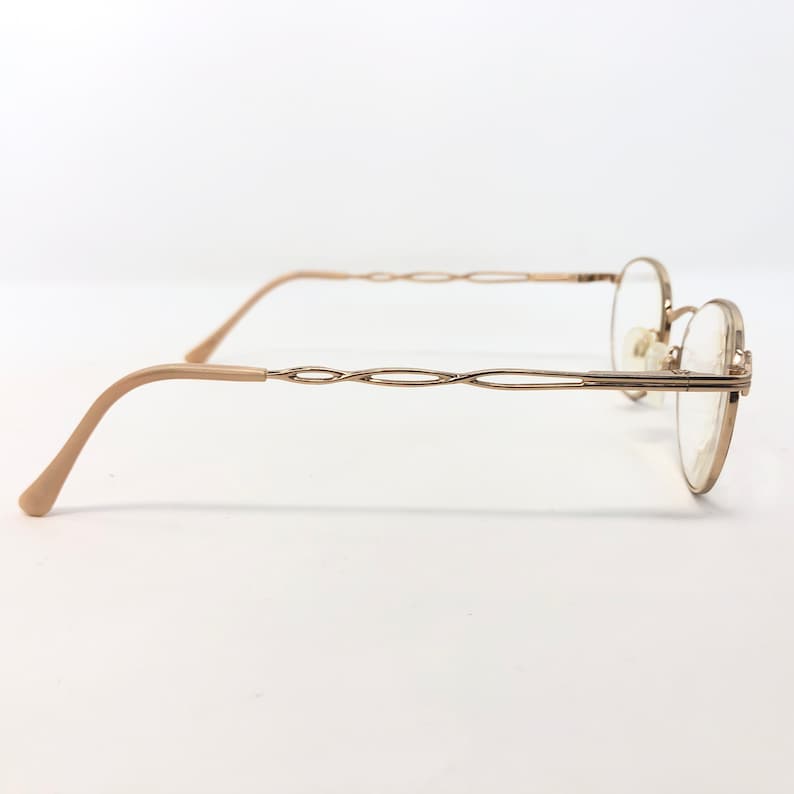 K&ouml;nnte beinhalten: Goldfarbene Brille mit zartem, ovalem Rahmen und einem einzigartigen, ineinander verschlungenen Design an den B&uuml;geln. Die B&uuml;gel und Ohrauflagen sind hellbeige. Die Gl&auml;ser sind klar, vor wei&szlig;em Hintergrund.