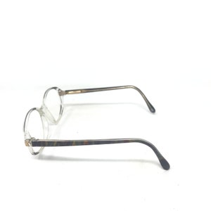 May include: Clear-framed spectacles with round lenses. The temples feature a marbled pattern in shades of brown, green, and grey. The arms are thin and curve down to the ear pieces. The spectacles are set against a white background.