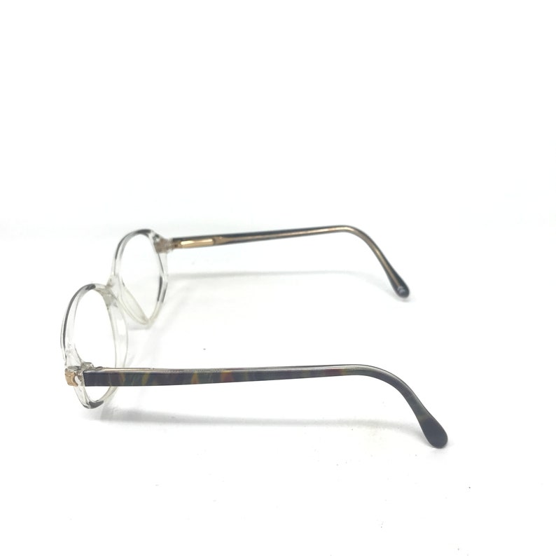 May include: Clear-framed spectacles with round lenses. The temples feature a marbled pattern in shades of brown, green, and grey. The arms are thin and curve down to the ear pieces. The spectacles are set against a white background.