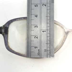 May include: Close-up of a pair of eyeglasses with a ruler measuring the lens. The lens is approximately 1 inch wide. The frame is a translucent gray color.